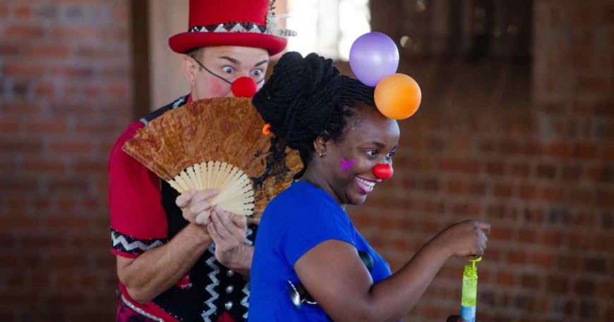 Two clowns perform, one smiles at the audience while the other hides behind her with a fan in front of his face.